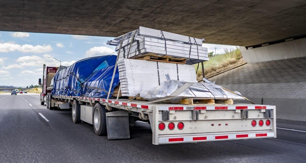 Hard loaded classic red big rig semi truck transporting covered and tightened commercial cargo on flat bed semi trailer running under the bridge across wide multiline highway road for delivery