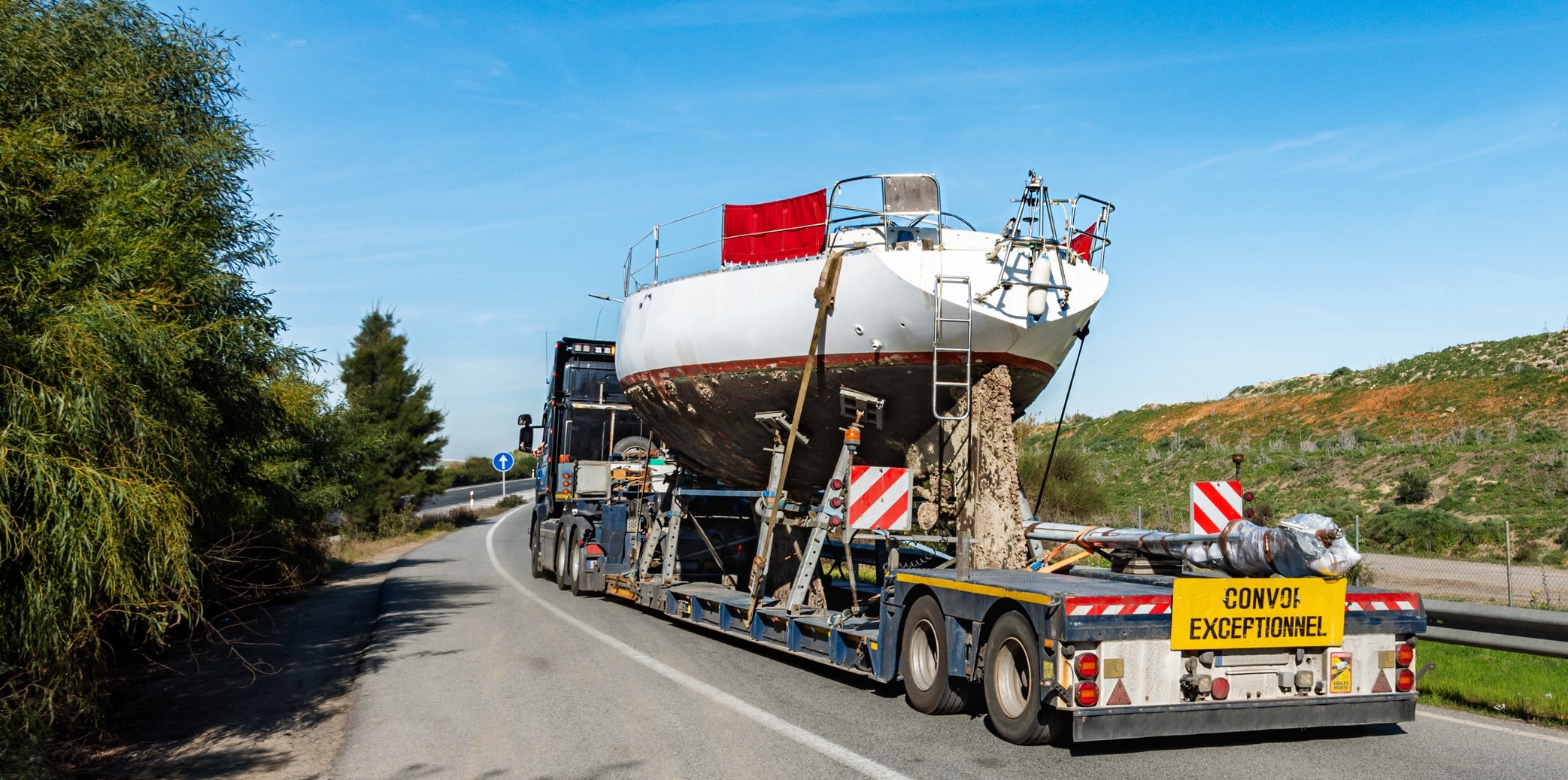 Truck with special semi-trailer for transporting boats carrying a sailboat on top.